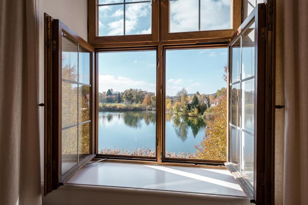 FAIRYTALE Castle Hotels in Bavaria 20 Open wooden-framed window with clear glass panes overlooks a calm lake surrounded by autumn foliage, under a blue sky. The peaceful scene evokes the tranquil charm found at castle hotels in Bavaria.