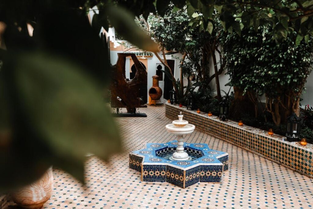 9 UNIQUE Riads in Tangier 33 A courtyard inspired by riads in Tangier, with patterned tile flooring, a small central fountain, and lush green trees. Decorative tiles line the raised edges, with lanterns along the side. Leaves in the foreground partly frame the view.