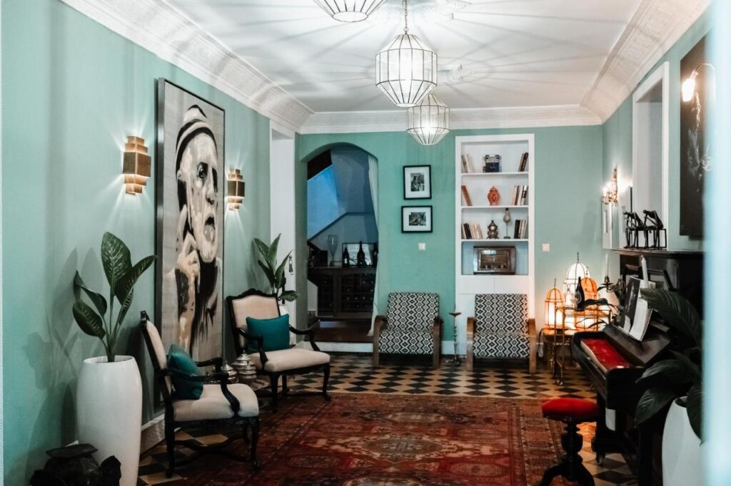 9 UNIQUE Riads in Tangier 32 A stylish living room inspired by riads in Tangier, with teal walls, eclectic furniture, plants, a piano, bookshelves, framed art, modern pendant lights, and a large portrait painting atop a patterned carpet and black-and-white tiled floor.