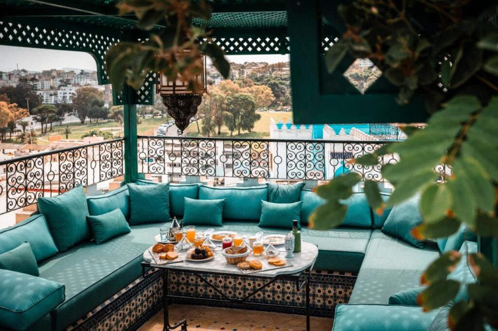 9 UNIQUE Riads in Tangier 31 A cozy outdoor lounge inspired by riads in Tangier features turquoise cushions around a round table set with breakfast, beneath a decorative canopy. Ornate railing and a city park are visible in the background.
