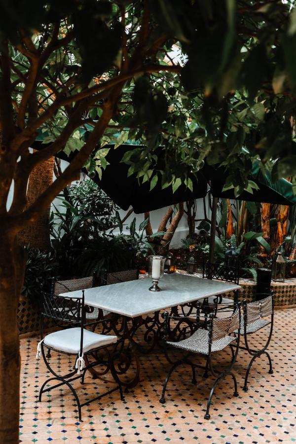 9 UNIQUE Riads in Tangier 30 A white marble table with four wrought iron chairs sits on a tiled patio surrounded by lush green plants and trees, evoking the cozy, shaded outdoor dining spaces found in traditional riads in Tangier.