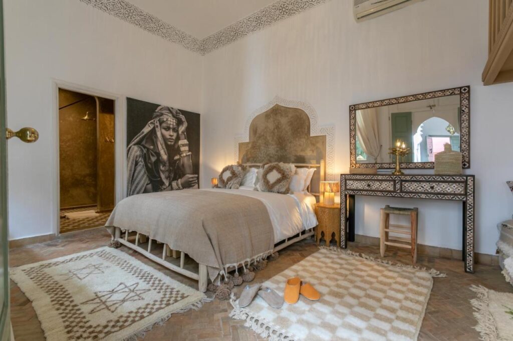 12 Charming Budget Riads in Marrakech 39 A cozy bedroom inspired by budget riads in Marrakech, featuring a beige and white color scheme, patterned rugs, a mirror and vanity, large wall art, candles beside the bed, textured walls, and ethnic-inspired decor.