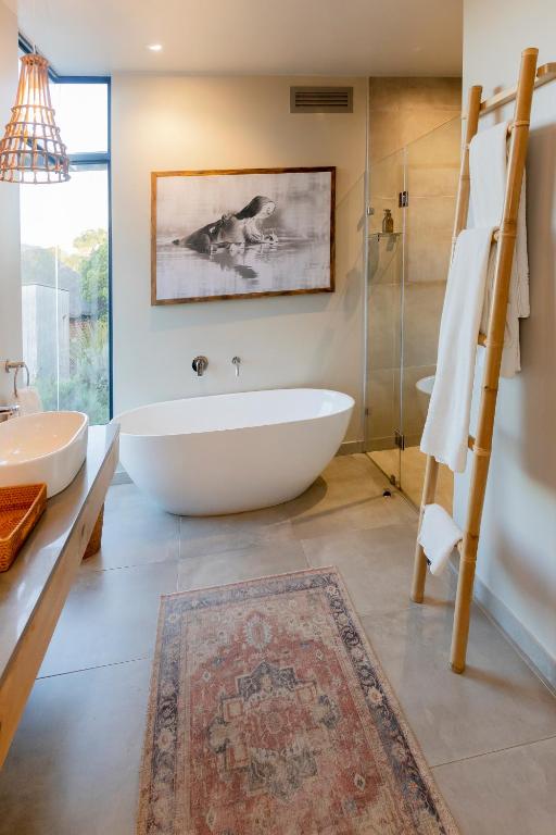 Modern bathroom inspired by Game Lodges in Western Cape, featuring a freestanding oval bathtub, glass shower, bamboo towel rack with white towels, woven light fixture, framed hippo photo, and patterned rug on gray tile floor.