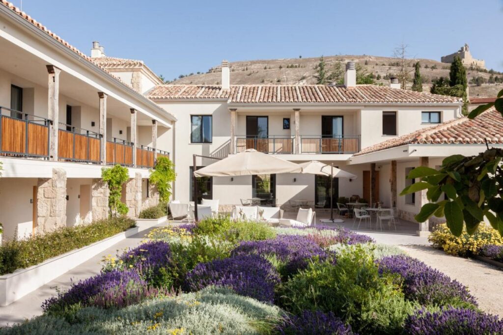 25 Weekend Trips from Madrid Max. 2h Away 62 A charming courtyard with blooming lavender and lush plants is surrounded by a two-story Mediterranean-style building. Perfect for weekend trips from Madrid, it features tiled roofs, balconies, and outdoor seating under umbrellas with a hillside and castle beyond.