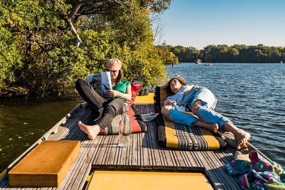 Boutique Hotels in Berlin Kreuzberg 42 Two people relax on a wooden dock by a lake, one reading a magazine and the other lying down with eyes closedādreaming perhaps of boutique hotels in Berlin Kreuzberg. Colorful mats and a bag are nearby, surrounded by trees and blue sky.