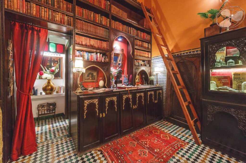 7 UNIQUE Riads in Fez You Won't Forget 22 A cozy, ornate library reminiscent of riads in Fez, with shelves of old books, a wooden ladder, patterned tile floor, red curtains and rug, plus a small room with a desk and lamp visible through an archway.
