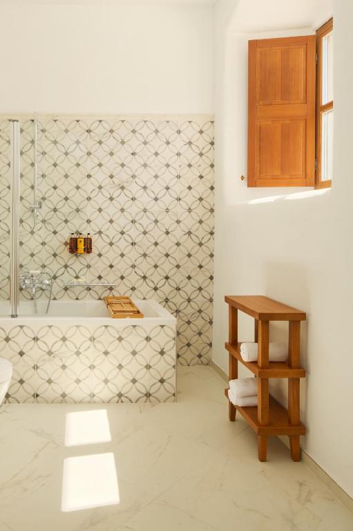 Most Unique Boutique Hotels in Dubrovnik 20 A bright bathroom reminiscent of boutique hotels in Dubrovnik, with patterned tile walls, a white bathtub, a wooden bath tray, and a small wooden shelf holding rolled towels. Sunlight pours in through a wooden window on the white wall.