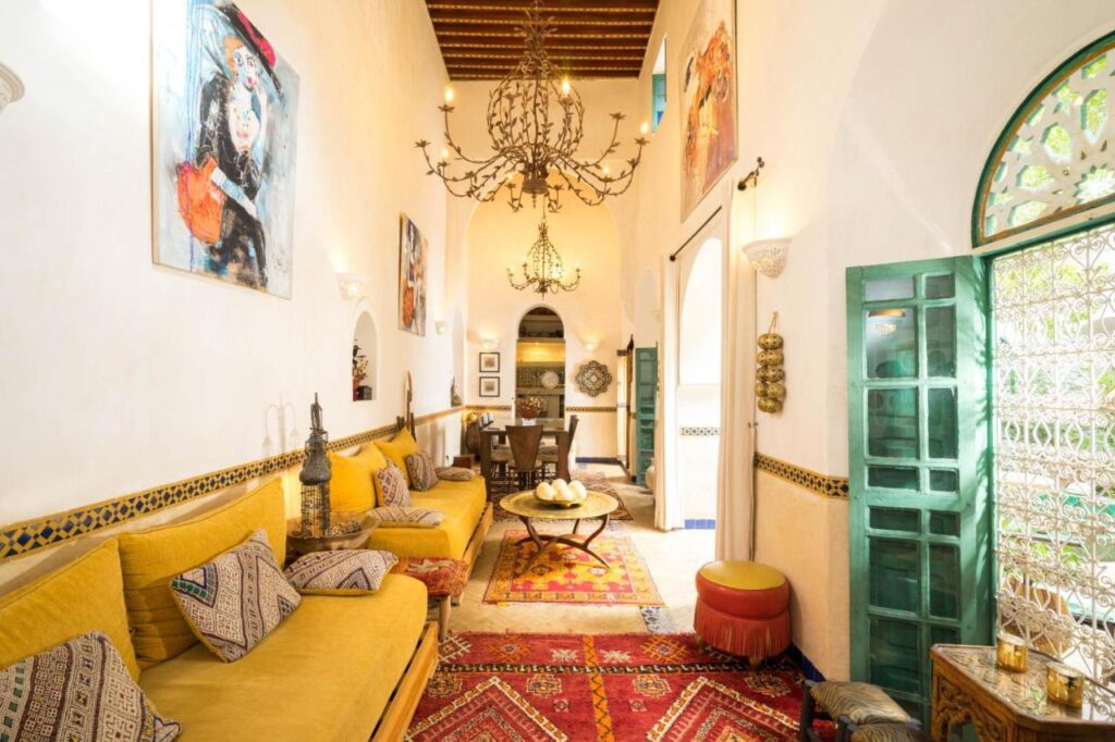 12 Charming Budget Riads in Marrakech 12 A bright, high-ceilinged living room with yellow sofas, patterned pillows, colorful rugs, ornate chandeliers, wall art, and green double doors opening to sunlight—reminiscent of budget riads in Marrakech with intricate Moroccan-inspired details.