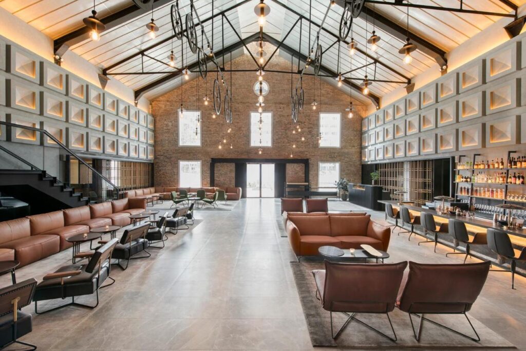 13 Most Unique Hotels in Singapore for an Incredible Holiday 17 Spacious modern lounge at one of the unique hotels in Singapore, featuring a high glass ceiling, brown leather sofas, armchairs, a bar area on the right, geometric wall panels, and large windows letting in natural light.
