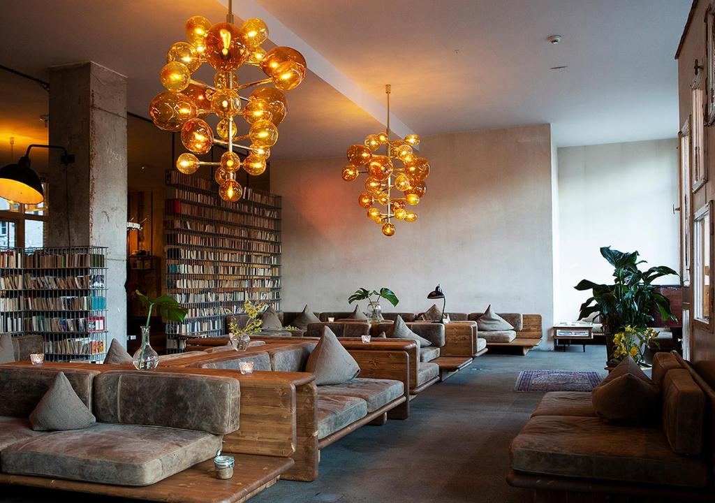 Boutique Hotels in Berlin Kreuzberg 8 A cozy lounge inspired by boutique hotels in Berlin Kreuzberg, with wooden sofas, brown cushions, low tables, large bookshelves, modern bubble chandeliers, potted plants, and warm ambient lighting in a spacious, minimalist room.