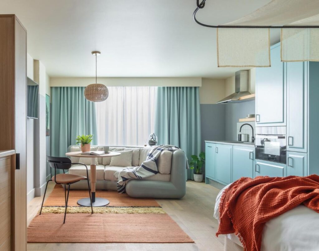 15 Best Boutique Hotels in London, UK 28 A modern studio apartment inspired by the best boutique hotels London offers, featuring light blue cabinets, a cozy kitchen, a round dining table for two, a beige sofa, an orange-accented bed, and large windows with light blue curtains.