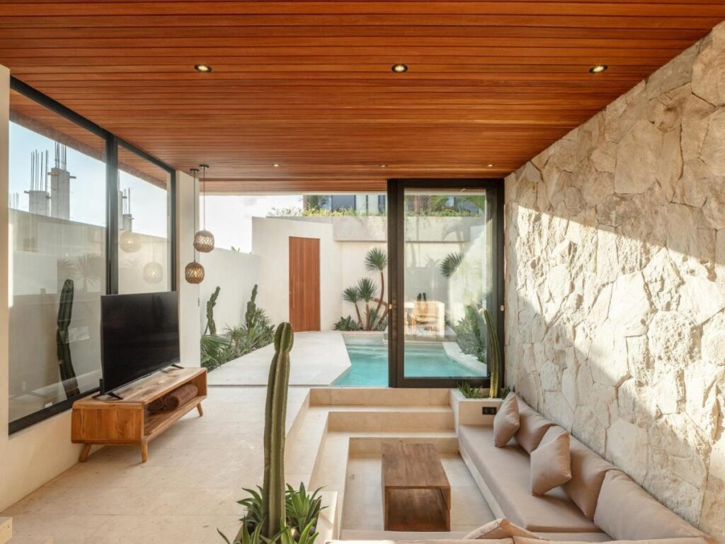 14 UNIQUE Boutique Hotels in Canggu, Bali 56 Modern living room with sunken seating, beige sofa, and a wooden coffee table, reminiscent of boutique hotels in Canggu. Enjoy seamless access to a private outdoor pool through glass doors, with a stone accent wall and minimalist décor completing the space.