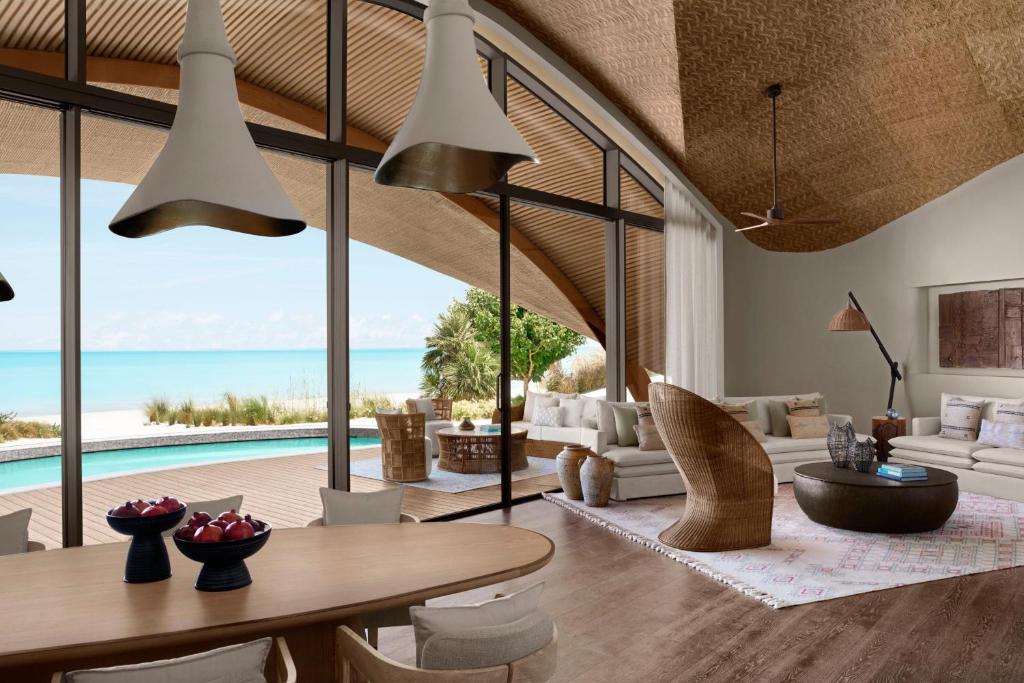 9 Best Hotels in Saudi Arabia: Unique Stays Across the Country 32 Modern living room with floor-to-ceiling windows overlooking a pool, palm trees, and a beach. Reminiscent of the Best Hotels in Saudi Arabia, it features woven furniture, neutral tones, a curved wooden ceiling, and a dining table with fresh fruit bowls.