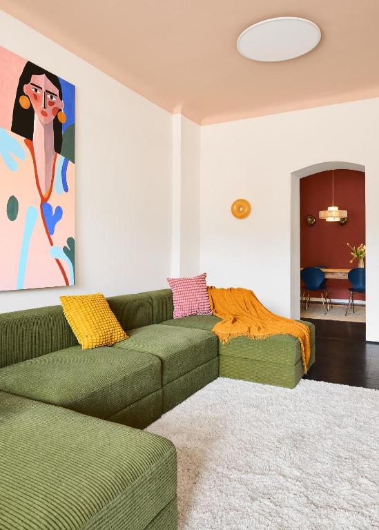 Boutique Hotels in Berlin Kreuzberg 22 A modern living room inspired by boutique hotels in Berlin Kreuzberg, featuring a green sectional sofa, yellow and red pillows, a mustard throw blanket, white rug, abstract portrait artwork, and a dining area visible through an arched doorway.