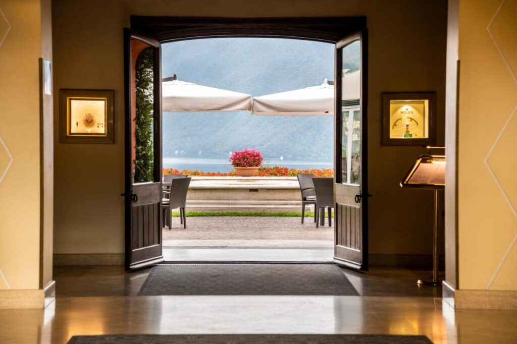 View through open double doors reveals a terrace with tables, chairs, large white umbrellas, and vibrant pink flowers in a planter—overlooking a lake and mountains at one of the best hotels in Lugano.