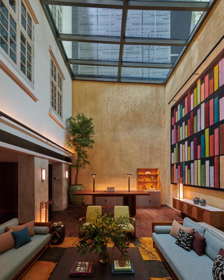 13 Most Unique Hotels in Singapore for an Incredible Holiday 33 A modern living room with a high ceiling and glass roof, colorful abstract wall art, cozy sofas, indoor plants, wooden floors, and large windows—evoking the vibrant style found in unique hotels in Singapore.