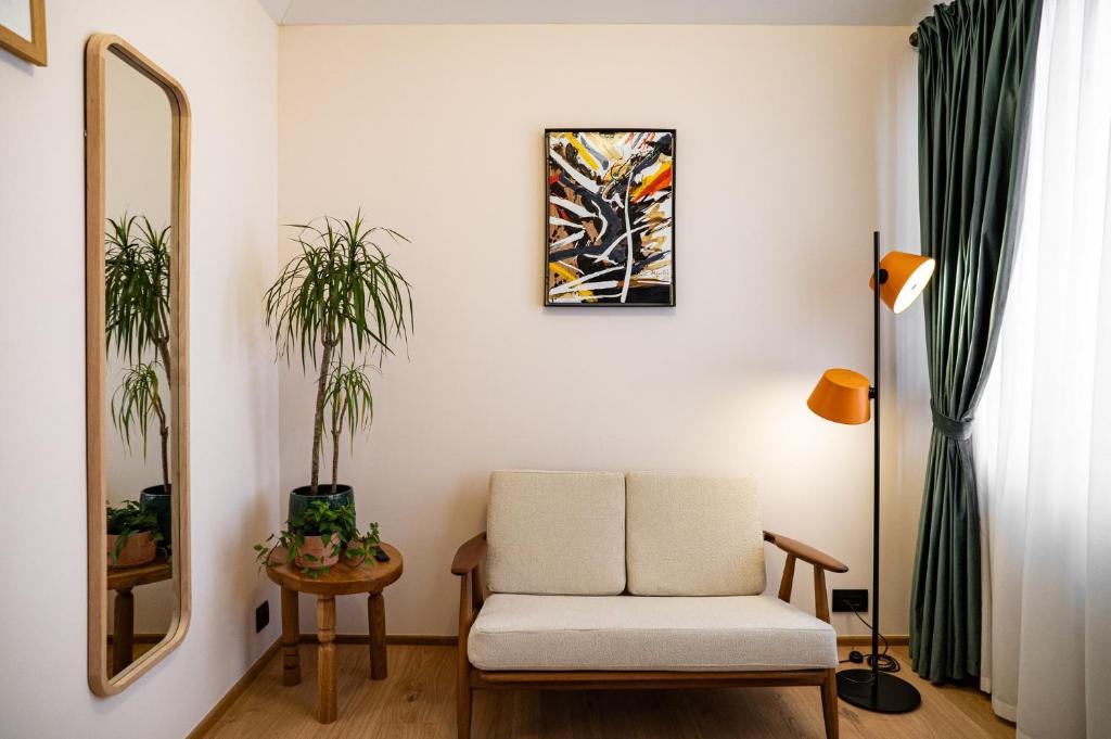 Most Unique Boutique Hotels in Dubrovnik 16 A cozy living room corner reminiscent of boutique hotels in Dubrovnik, with a light wood loveseat, beige cushions, a potted plant on a round wooden side table, a tall floor lamp with orange shades, green curtains, and abstract art on a pale wall.