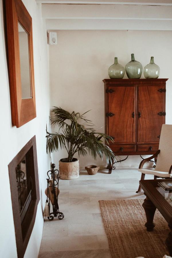 Unique Agroturismos in Mallorca 29 A cozy living room inspired by agroturismos in Mallorca, featuring a wooden cabinet topped with three green glass bottles, a potted palm plant, a fireplace, a woven rug, and neutral-toned furniture. The decor is simple and elegant.