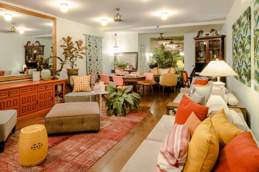 13 Most Unique Hotels in Singapore for an Incredible Holiday 44 A cozy, eclectic living and dining room inspired by unique hotels in Singapore, with colorful pillows, plants, a large mirror, patterned rugs, a red sideboard, gray ottoman, and a wooden dining table with pink chairs under warm lighting.