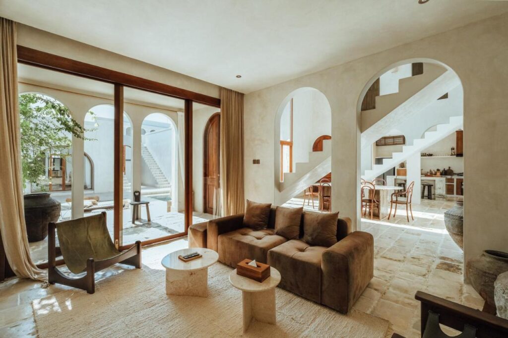 14 UNIQUE Boutique Hotels in Canggu, Bali 16 A sunlit living room with a brown sofa, rocking chair, and round tables evokes the inviting charm of boutique hotels in Canggu, featuring arched doorways, stone floors, and an open view into a bright kitchen and staircase.