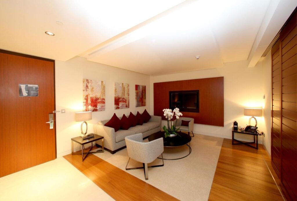 Where to Stay in Doha, Qatar: Transit, City & Beach Hotels 23 Modern hotel suite living area with a gray sofa, armchairs, coffee table featuring white orchids—an inviting choice when searching for where to stay in Doha. Enjoy wall art, a flat-screen TV, wood accents, and warm lighting from two lamps on side tables.