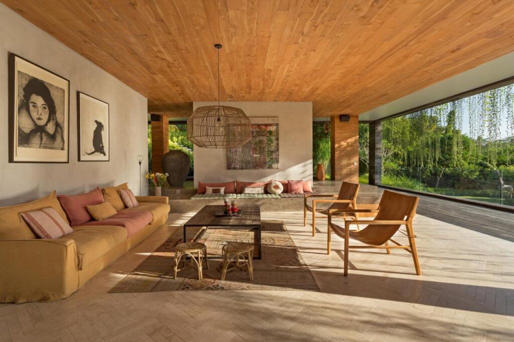 A modern living room with large windows, wood ceiling, tan sofa, minimalist chairs, artwork on the wall, and a woven pendant light offers views of greenery—reminiscent of the best family villas in Bali. Natural light fills this cozy, stylish space.
