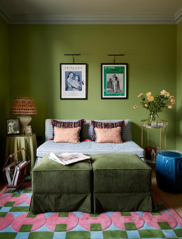 15 Best Boutique Hotels in London, UK 47 A colorful living room inspired by boutique hotels in London, with green walls, a blue patterned sofa with pink cushions, two framed artworks above, a green corduroy ottoman, geometric rug, flowers on a gold side table, and a blue stool.