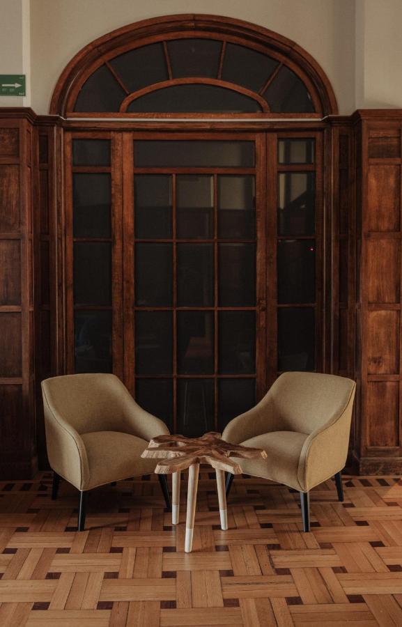 Best Boutique Hotels in San Sebastian 31 Two beige armchairs face each other with a small wooden table between them, set on a patterned wooden floor—creating an inviting scene reminiscent of boutique hotels in San Sebastian, framed by tall, dark wood-framed glass doors and paneled walls.