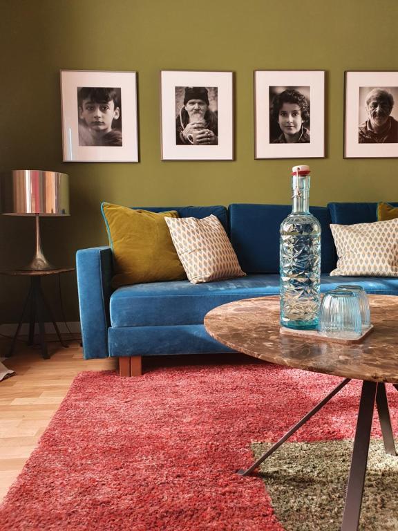 Unique Boutique Hotels in Salzburg 27 A cozy living room, inspired by boutique hotels in Salzburg, features a blue sofa with patterned cushions and a red rug. A round coffee table holds a clear bottle and glass. Four black-and-white portraits hang on an olive-green wall behind the sofa.