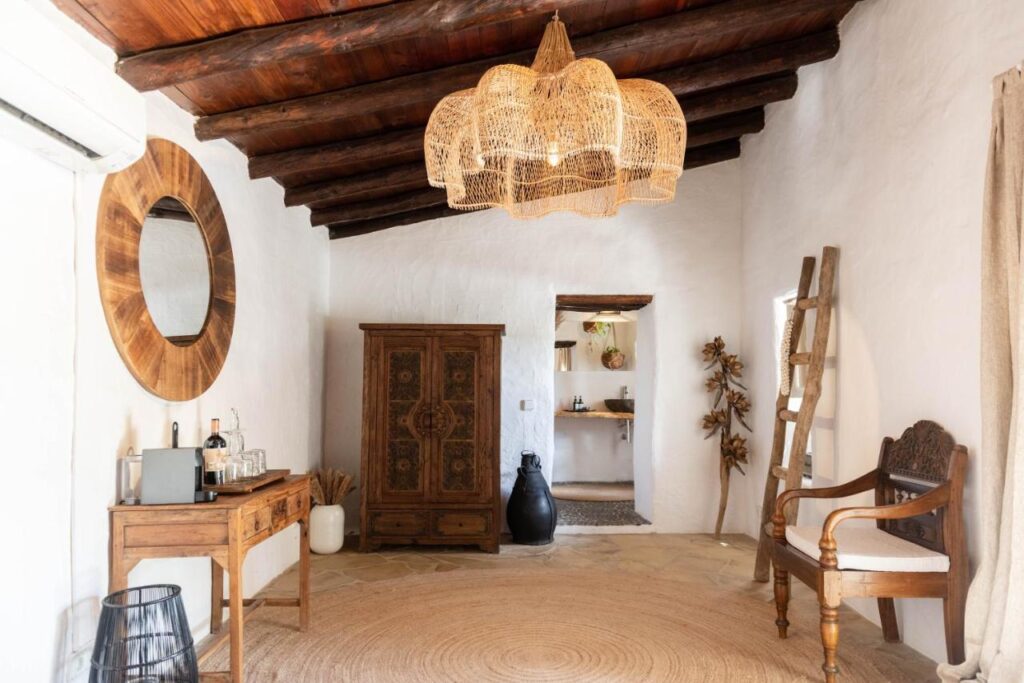 10 AUTHENTIC Finca Hotels in Ibiza 9 A rustic room in the style of finca hotels in Ibiza, with white plaster walls, wooden ceiling beams, a wicker chandelier, a round mirror above a small desk, a vintage wardrobe, a ladder, and a carved chair on a jute rug.