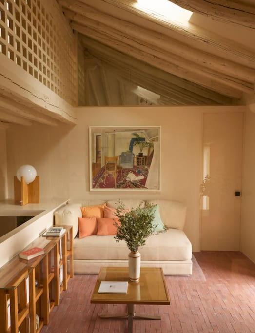 25 Weekend Trips from Madrid Max. 2h Away 60 Cozy living room with slanted wooden ceiling, cream walls, a white sofa with pastel cushions, and a modern lamp—perfect for planning weekend trips from Madrid. A wooden coffee table with a plant and colorful artwork complete the bright, inviting space.