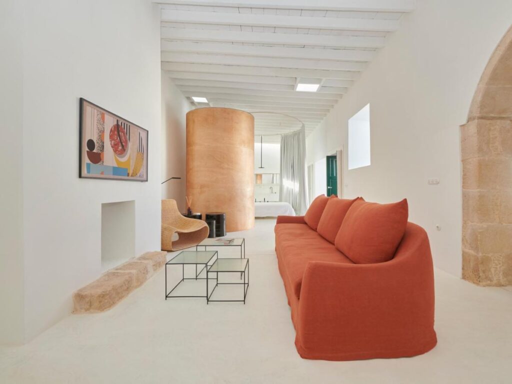 Adults Only Hotels in Fuerteventura 3 Minimalist living space with white walls, a large orange sofa, glass coffee tables, abstract wall art, and a wicker chair. Reminiscent of stylish adults only hotels in Fuerteventura, the room features a tan curved accent wall and abundant natural light.