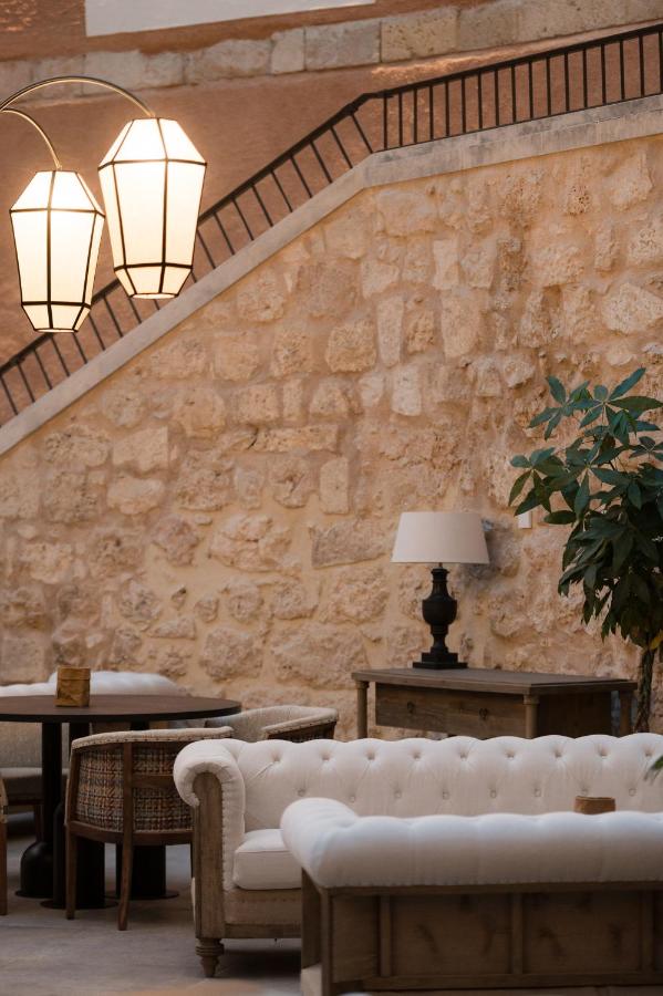 25 Weekend Trips from Madrid Max. 2h Away 95 A cozy indoor seating area with cream-colored tufted sofas, wooden tables, a lamp on a side table, potted plant, and a stone wall—perfect for planning weekend trips from Madrid—with a staircase and large hanging lanterns in the background.