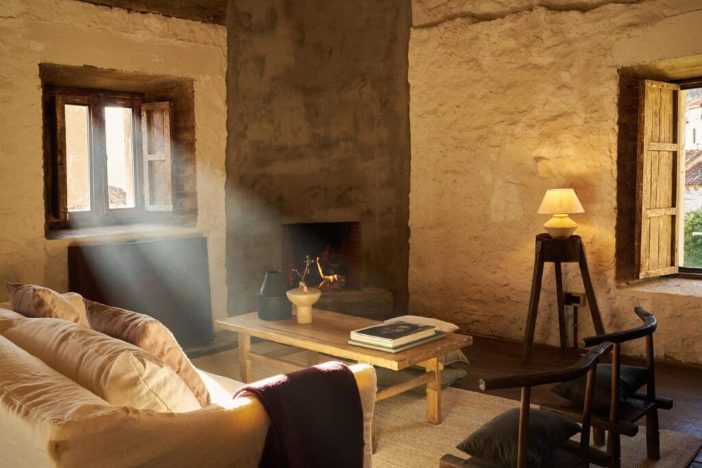 Sunlight streams through rustic wooden windows into a cozy room reminiscent of boutique hotels in Asturias, with textured stone walls, a lit fireplace, a beige sofa, a wooden coffee table stacked with books, and a table lamp creating an inviting atmosphere.
