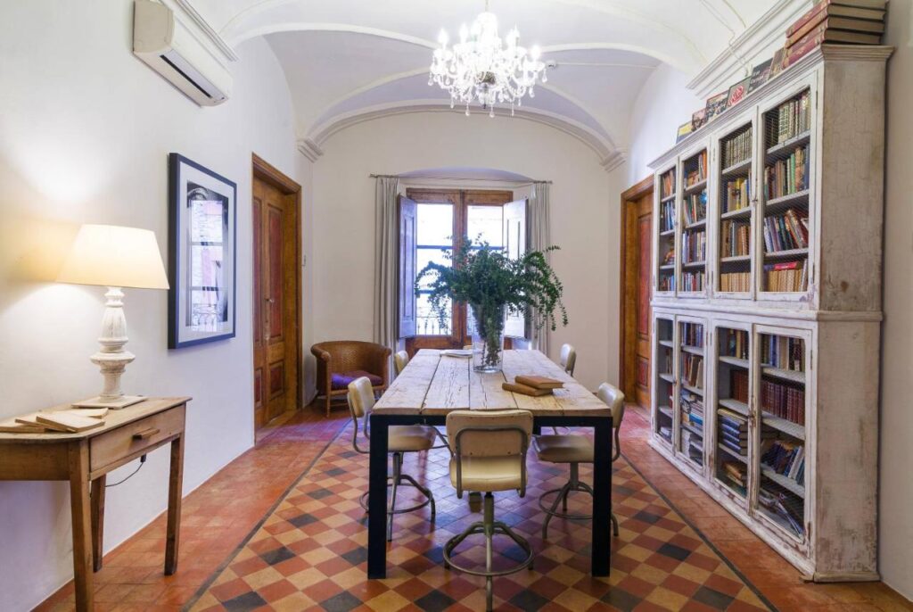 A bright, elegant dining room reminiscent of boutique hotels in Costa Brava, with a rustic wooden table, five chairs, a large bookshelf full of books, a chandelier, potted plant centerpiece, lamp on a side table, and a curtained window at the far end.