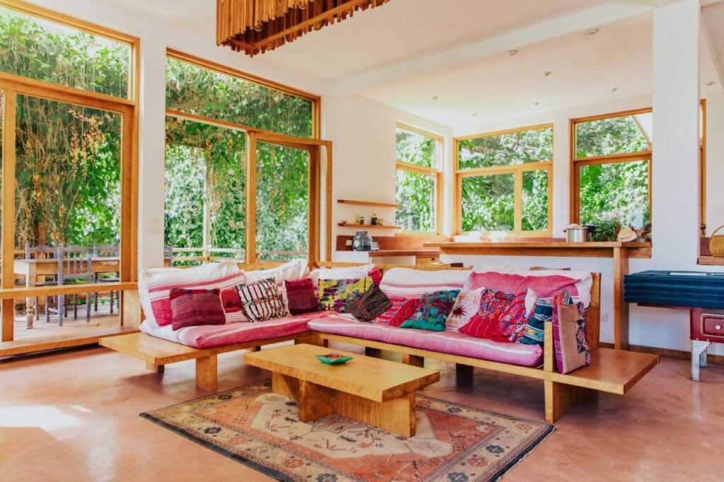 8 UNIQUE Places Where to Stay in Lake Atitlán 11 A bright living room with large windows, wooden furniture, and a colorful sofa with patterned cushions—perfect for those wondering where to stay in Lake Atitlan. Sunlight streams in, revealing lush greenery outside and a cozy, rustic-modern vibe.