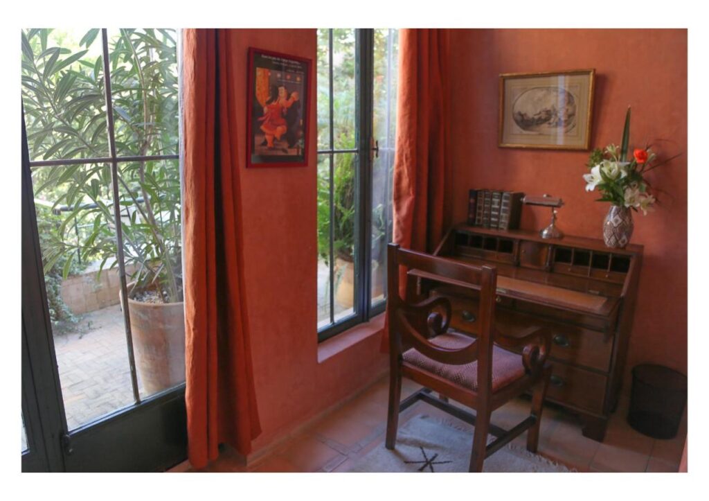 9 UNIQUE Riads in Tangier 11 A small home office with a wooden desk and chair by tall windows, orange walls, a vase of flowers, framed art, and books. Sunlight and greenery outside evoke the warm atmosphere found in riads in Tangier.