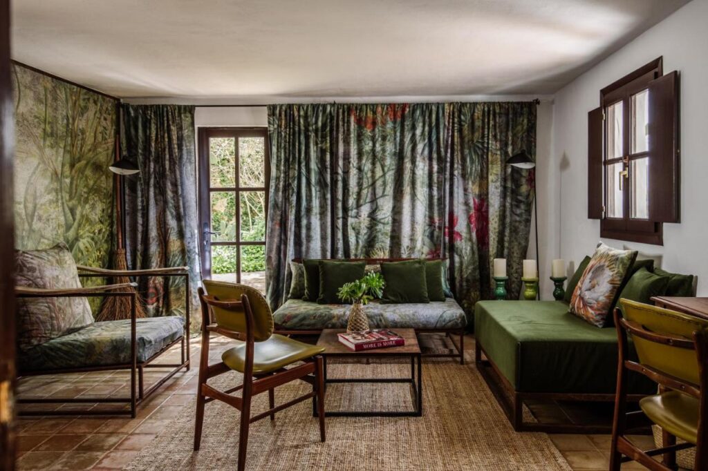 10 AUTHENTIC Finca Hotels in Ibiza 12 A cozy living room inspired by finca hotels in Ibiza, with green-themed decor, forest-patterned curtains and cushions, green sofas, wooden chairs, a woven rug, and a coffee table with books and a vase of leaves. Sunlight streams through the window.