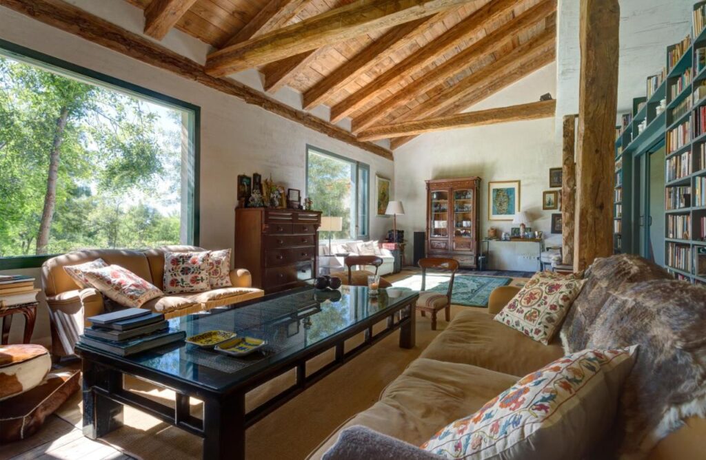 25 Weekend Trips from Madrid Max. 2h Away 50 A cozy living room with wooden beams, large windows, bookshelves, and comfortable sofas with patterned pillows—perfect for planning weekend trips from Madrid as sunlight streams in, creating a warm and inviting atmosphere.