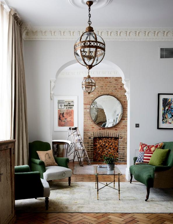 15 Best Boutique Hotels in London, UK 37 Eclectic living room with green armchairs, patterned pillows, exposed brick fireplace with stacked logs, round mirror, vintage posters, and pendant lights—reminiscent of the unique charm found in boutique hotels in London. Natural light filters through long curtains.