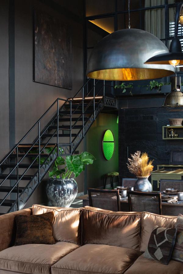 14 UNIQUE Boutique Hotels in Canggu, Bali 4 A modern living space inspired by boutique hotels in Canggu, featuring a brown velvet sofa, metal dome light over the dining table, dark walls, black-railed staircase, lush green plants, and a glowing green oval wall feature.