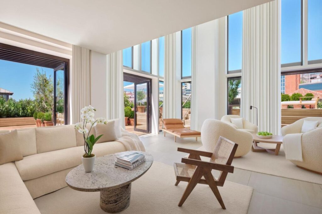 Where to Stay in Madrid: 14 Best Hotels 21 A bright, modern living room with high ceilings, floor-to-ceiling windows, cream-colored furniture, a round stone coffee table, and lush indoor plants—an ideal spot for those wondering where to stay in Madrid first time.