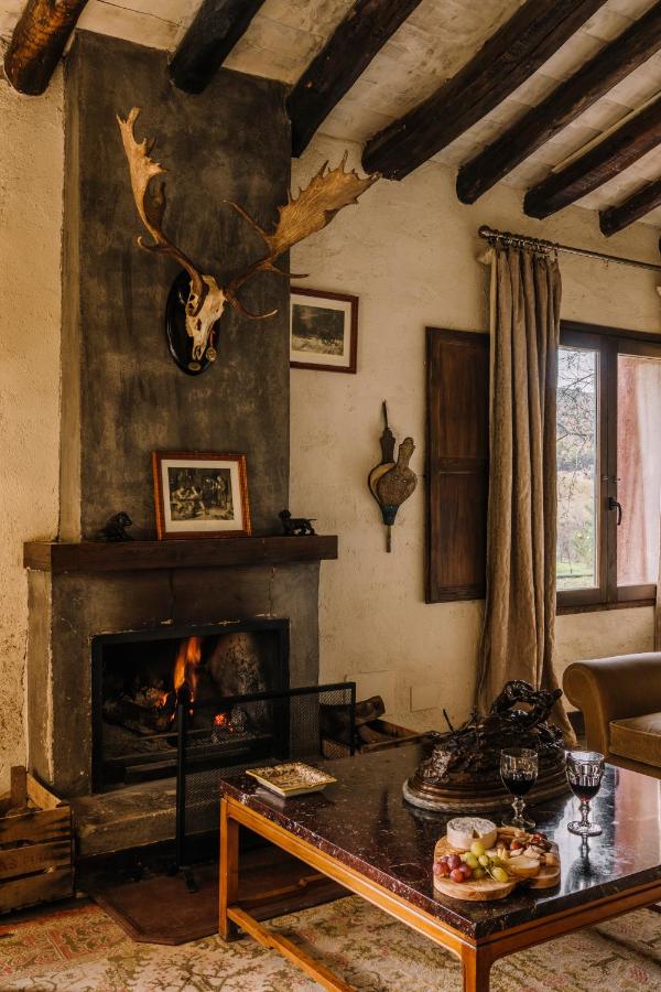 25 Weekend Trips from Madrid Max. 2h Away 46 A cozy rustic living room, perfect for weekend trips from Madrid, features a lit fireplace with antlers above it, framed art, and a coffee table set with wine, cheese, and fruit. Wooden beams line the ceiling as sunlight pours through draped windows.