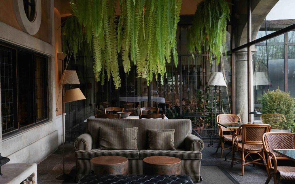25 Weekend Trips from Madrid Max. 2h Away 57 A cozy indoor lounge area, perfect for planning weekend trips from Madrid, features a striped sofa, round ottomans, wicker chairs, hanging ferns, floor lamps, and large glass windows overlooking a garden.