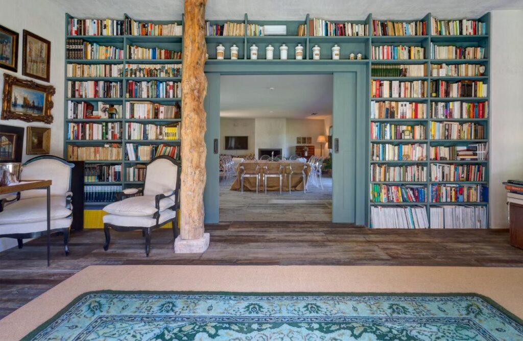25 Weekend Trips from Madrid Max. 2h Away 53 A cozy library room with bookshelves framing a doorway, two elegant armchairs, wooden floors, framed art on the wall, and a patterned rug—perfect for planning weekend trips from Madrid. The doorway leads to a bright living area with more seating.