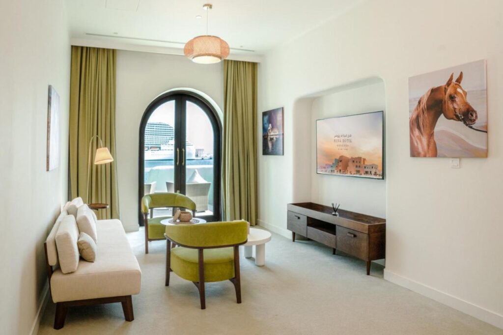 Where to Stay in Doha, Qatar: Transit, City & Beach Hotels 33 A modern living room with a beige sofa, two green chairs, a coffee table, TV on a wooden stand, wall art, and large arched windows with green curtains—ideal inspiration for those searching where to stay in Doha.