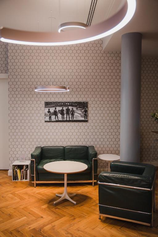 Authentic Boutique Hotels in Munich 27 A modern lounge area reminiscent of boutique hotels in Munich, featuring green armchairs, a round white table, a side table with magazines, hexagonal patterned wallpaper, and a black-and-white photo under circular ceiling lights.