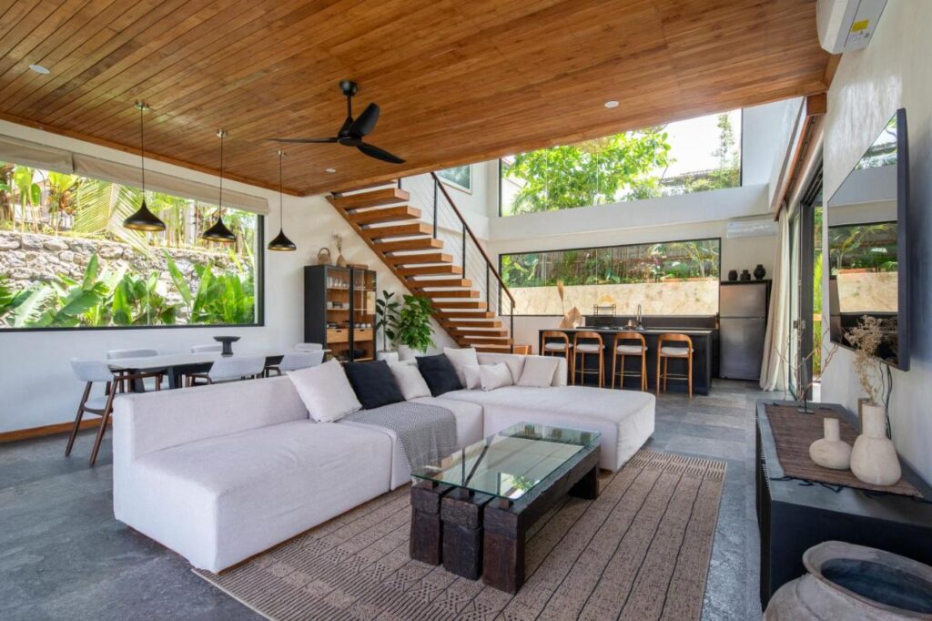 Modern open-concept living space with a white sectional sofa, glass coffee table, dining area, and kitchen. Large windows showcase lush greenery outside. Wooden ceiling and staircase add warmth to one of the best family villas in Bali.