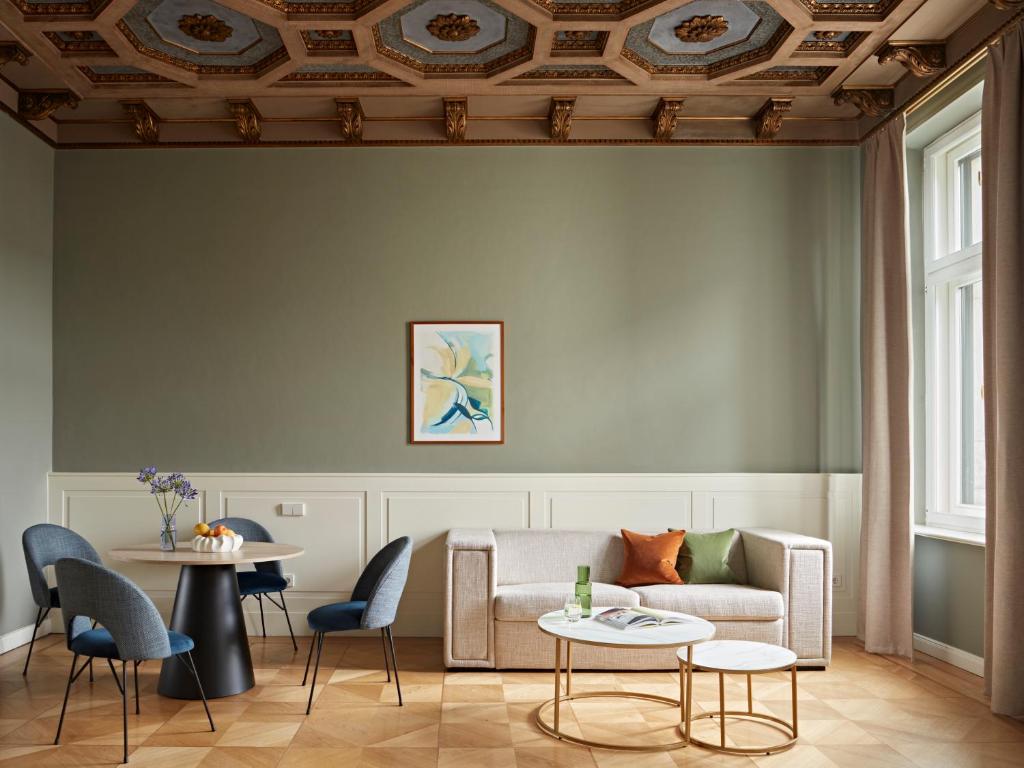 Boutique Hotels in Berlin Kreuzberg 18 A stylish living room inspired by boutique hotels in Berlin Kreuzberg, with a beige sofa, colorful cushions, round coffee tables, blue chairs around a black dining table, light green walls, and an ornate coffered ceiling with gold accents. Natural light pours in.