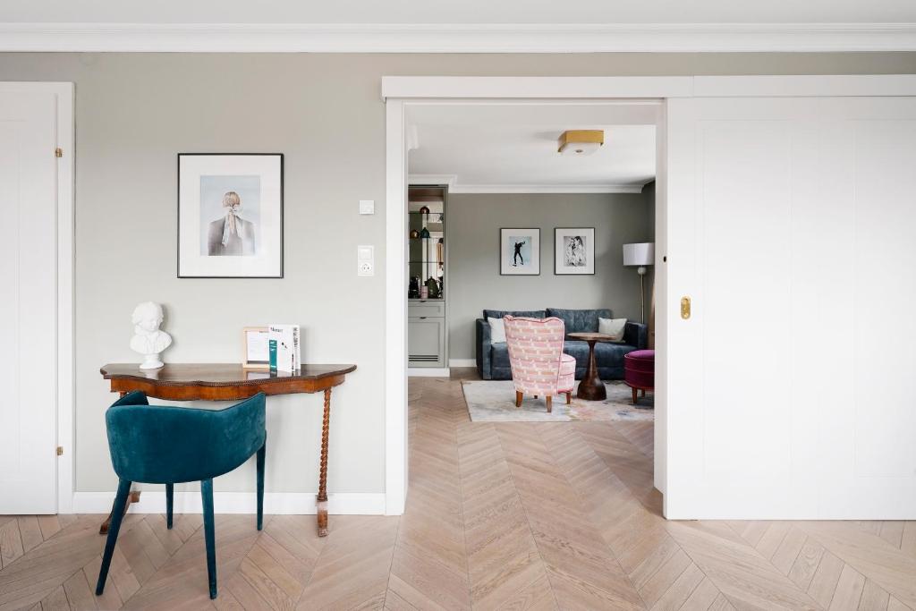 Unique Boutique Hotels in Salzburg 16 A stylish interior reminiscent of boutique hotels in Salzburg features a small wooden desk and green chair against a gray wall, leading into a cozy living room with a blue sofa, patterned armchair, framed art, and light wood herringbone floors throughout.